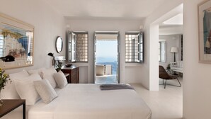 A spacious bedroom with a large bed, a desk, and a chair. There are multiple windows providing a view of the outdoors. - Ikia Kriton (Santorini)