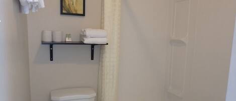 A bathroom with a toilet, a bathtub, a shower curtain, and a shelf with towels and toiletries.