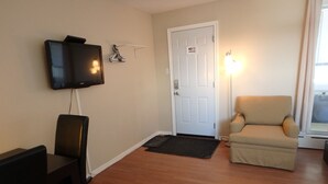 A living room with a flat-screen TV mounted on the wall, a door with a "No Smoking" sign, a cushioned armchair, and a small table with a chair. - Wheel Inn Motel (Temiskaming Shores)