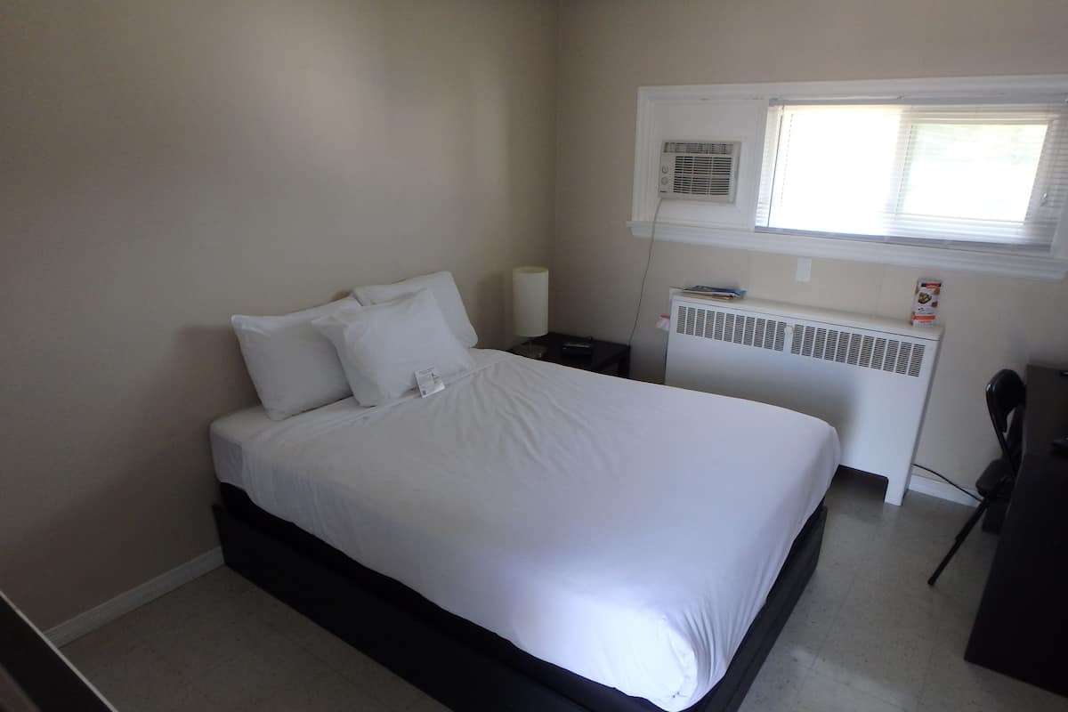 A neatly made bed in a hotel room with a white headboard, a bedside table with a lamp, a radiator, and a window with blinds.