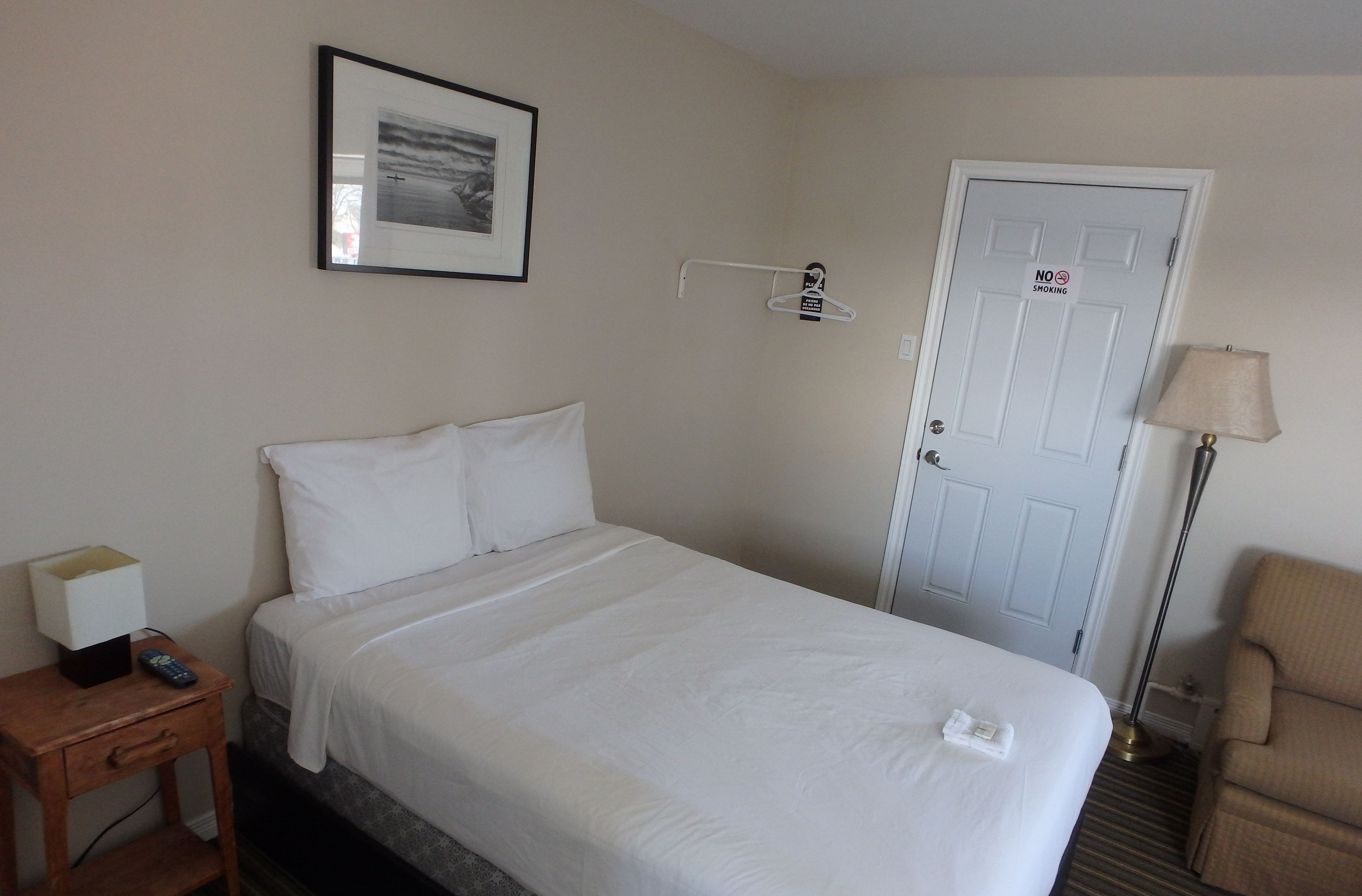 A hotel room with a bed, a nightstand with a lamp, a chair, and a door with a 'No Smoking' sign.