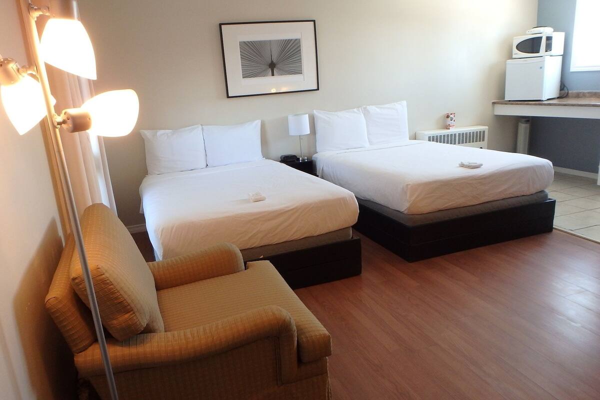 A hotel room with two beds, a chair, a desk with a microwave, and a framed picture on the wall.