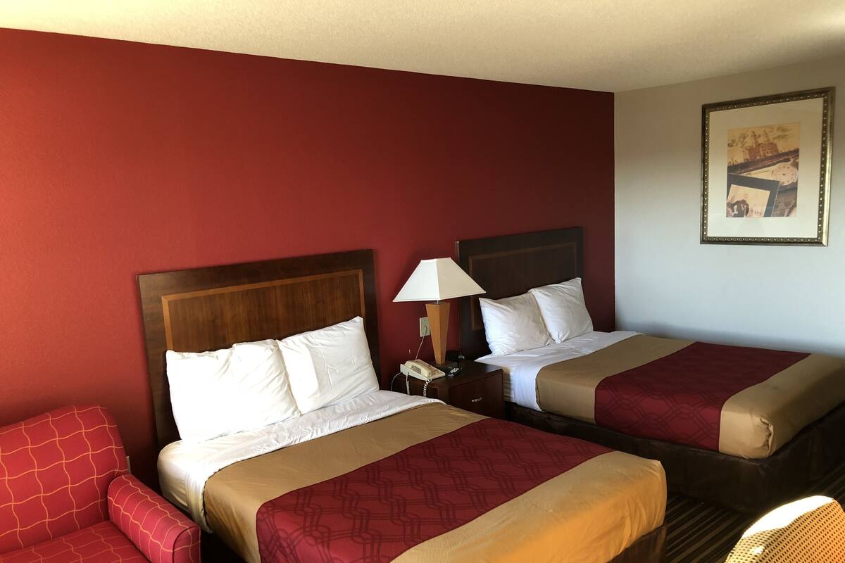 Quarto de hotel com duas camas, uma poltrona vermelha, mesa de cabeceira com abajur e um quadro na parede.