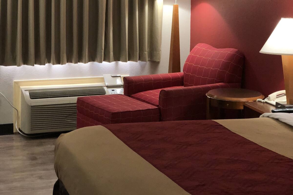 Quarto de hotel com uma cama, um sofá vermelho, uma escrivaninha com luminária, um telefone e uma janela com cortinas.