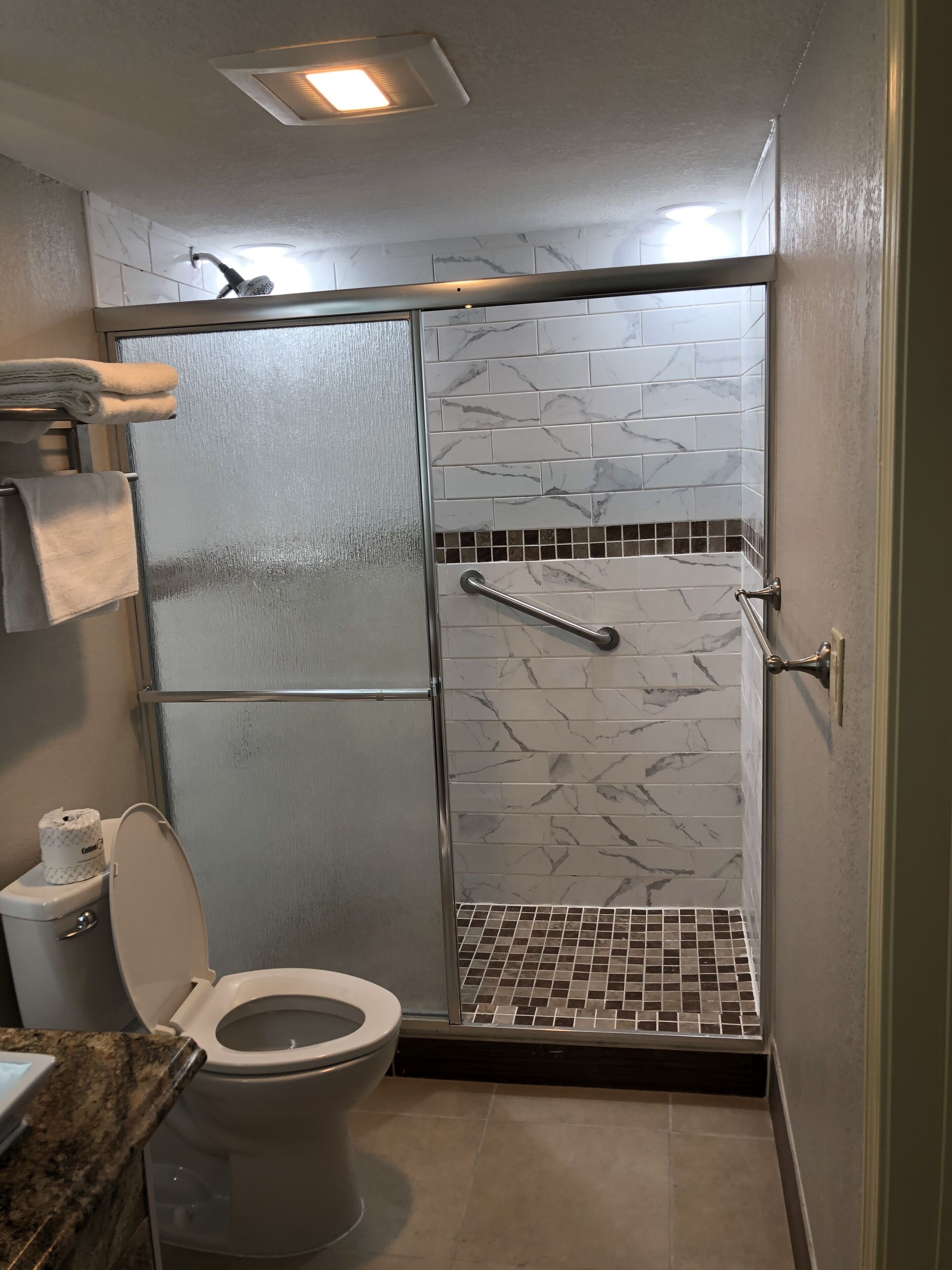 Ein Badezimmer mit Toilette, einer Dusche mit Glastür, Marmorfliesen und einer Haltestange.