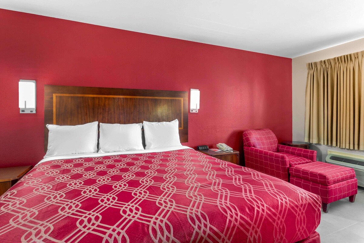 A hotel room with a large bed, a wooden headboard, a red patterned bedspread, a red armchair, a small table with a phone, and a window with beige curtains.