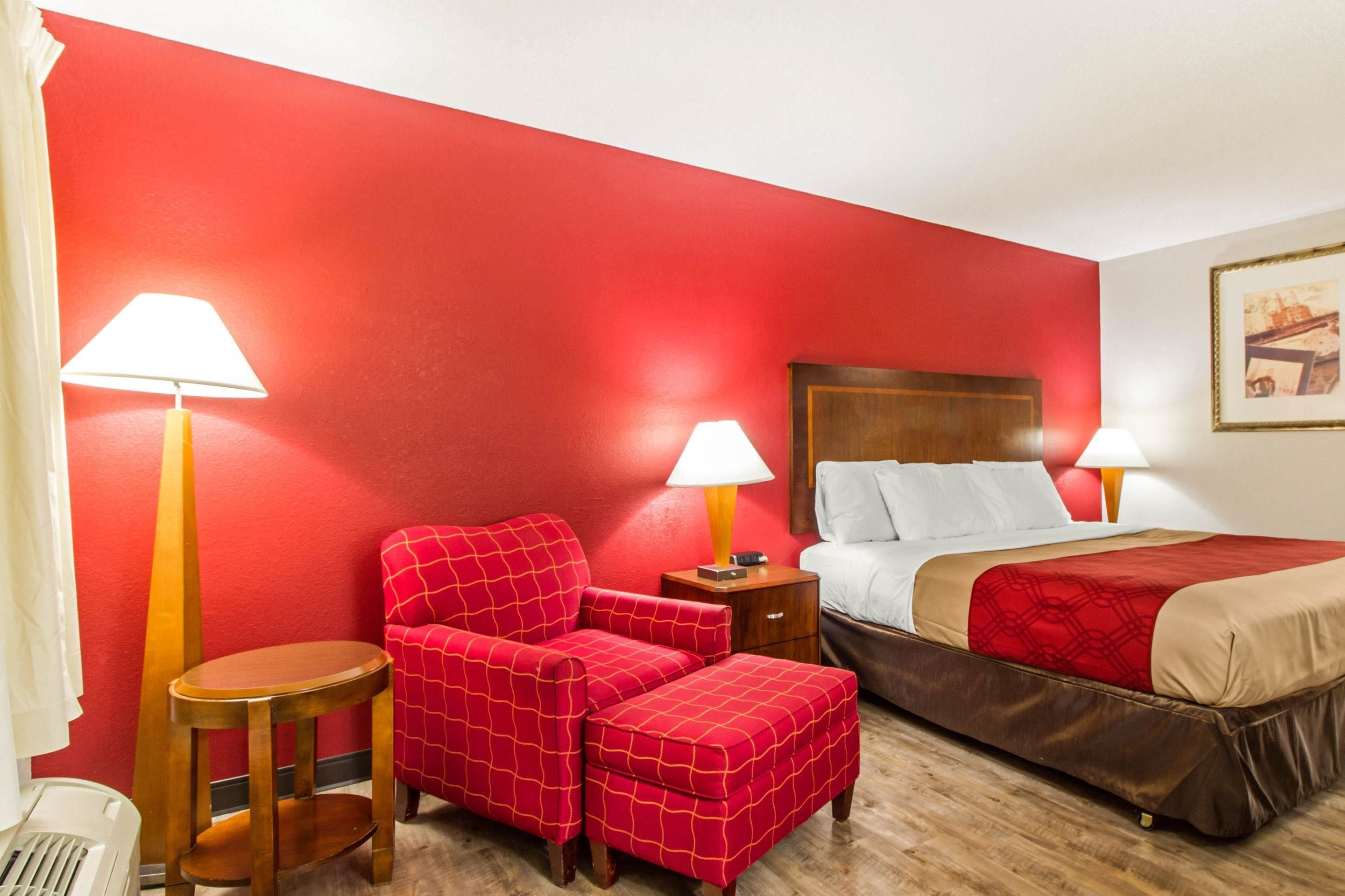 Habitación de hotel con pared roja, cama con cabecera de madera, dos lámparas de noche, un sillón rojo, una mesita de madera y un radiador blanco.