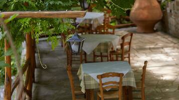 Een terras buiten met houten tafels en stoelen, bedekt met witte tafelkleden, omgeven door groen en een stenen vloer.