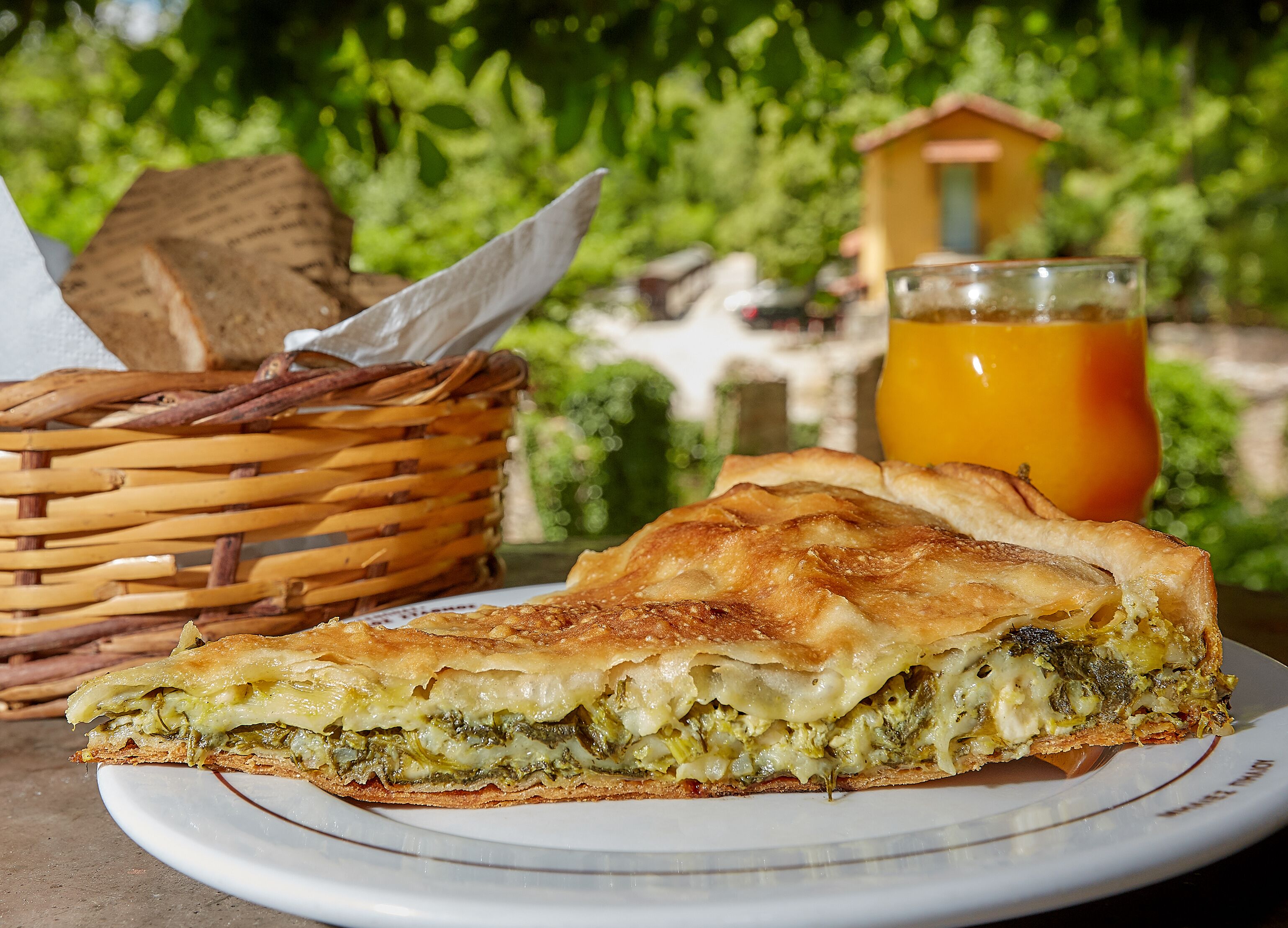 Een plakje quiche op een bord, met een mandje en een glas sinaasappelsap op de achtergrond.