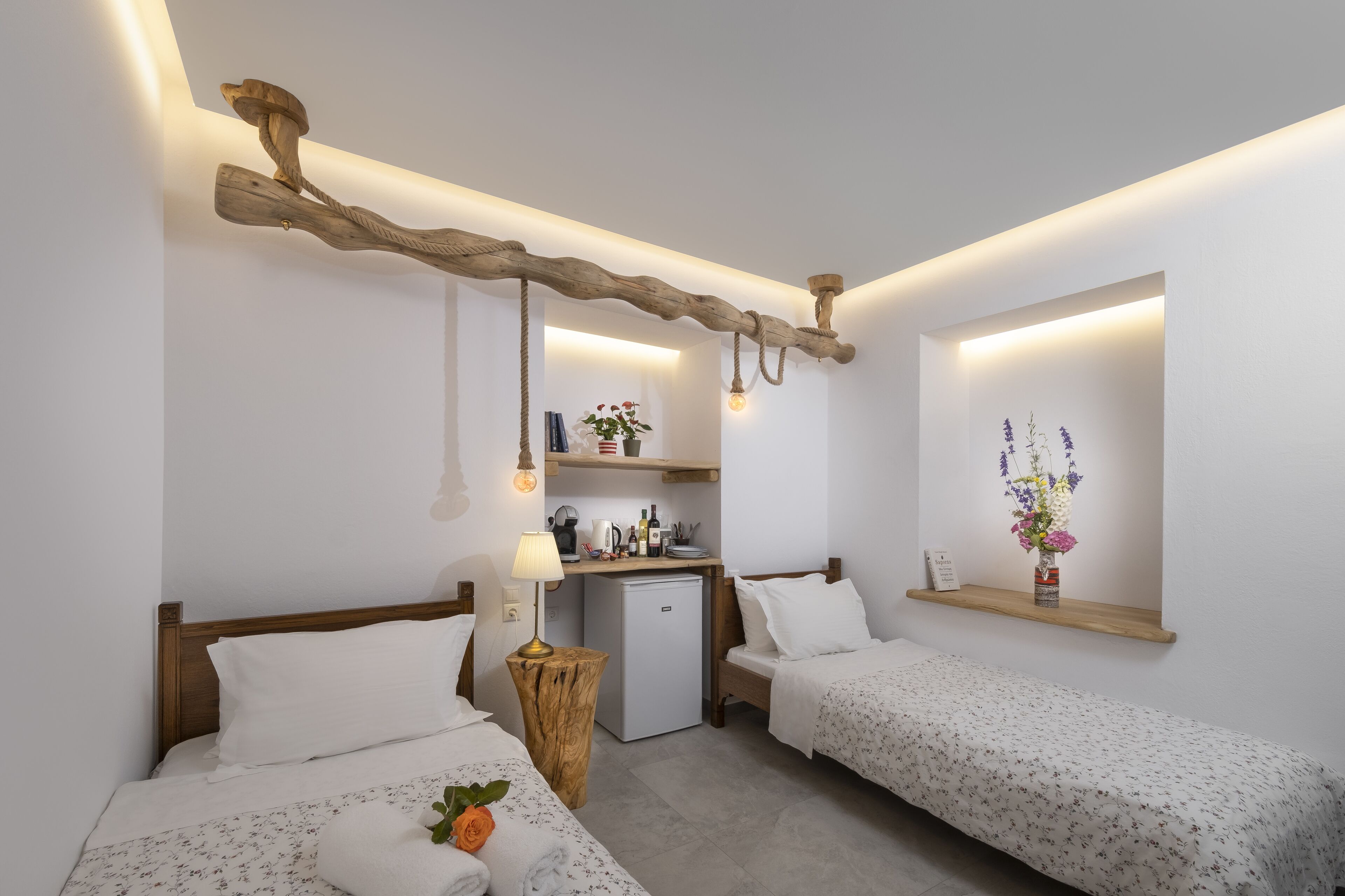 Quarto de hotel com duas camas, uma kitchenette pequena, uma estante de madeira com flores e um design rústico no teto.