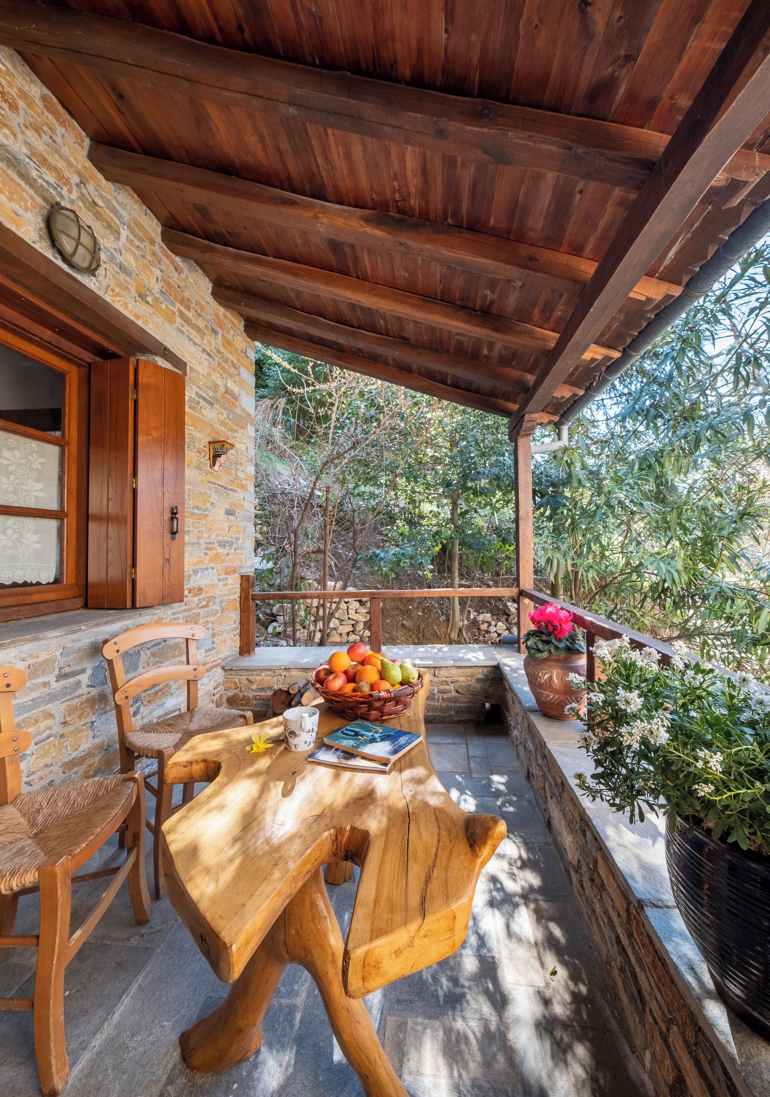 A wooden table with chairs and a fruit basket on a stone patio under a wooden pergola.