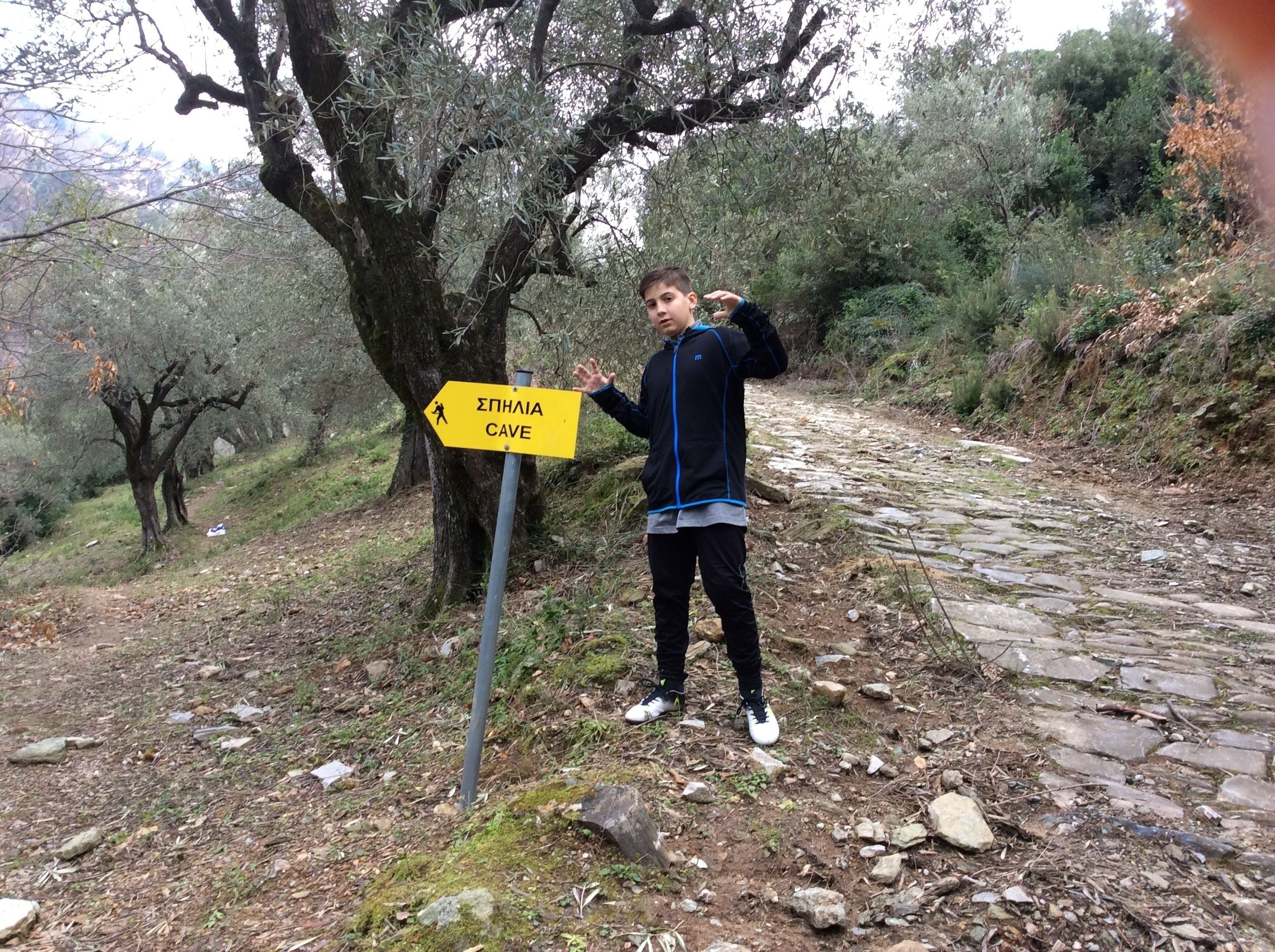 Una persona de pie junto a un cartel que indica 'Epinalia Cave' en un camino de piedra rodeado de árboles.