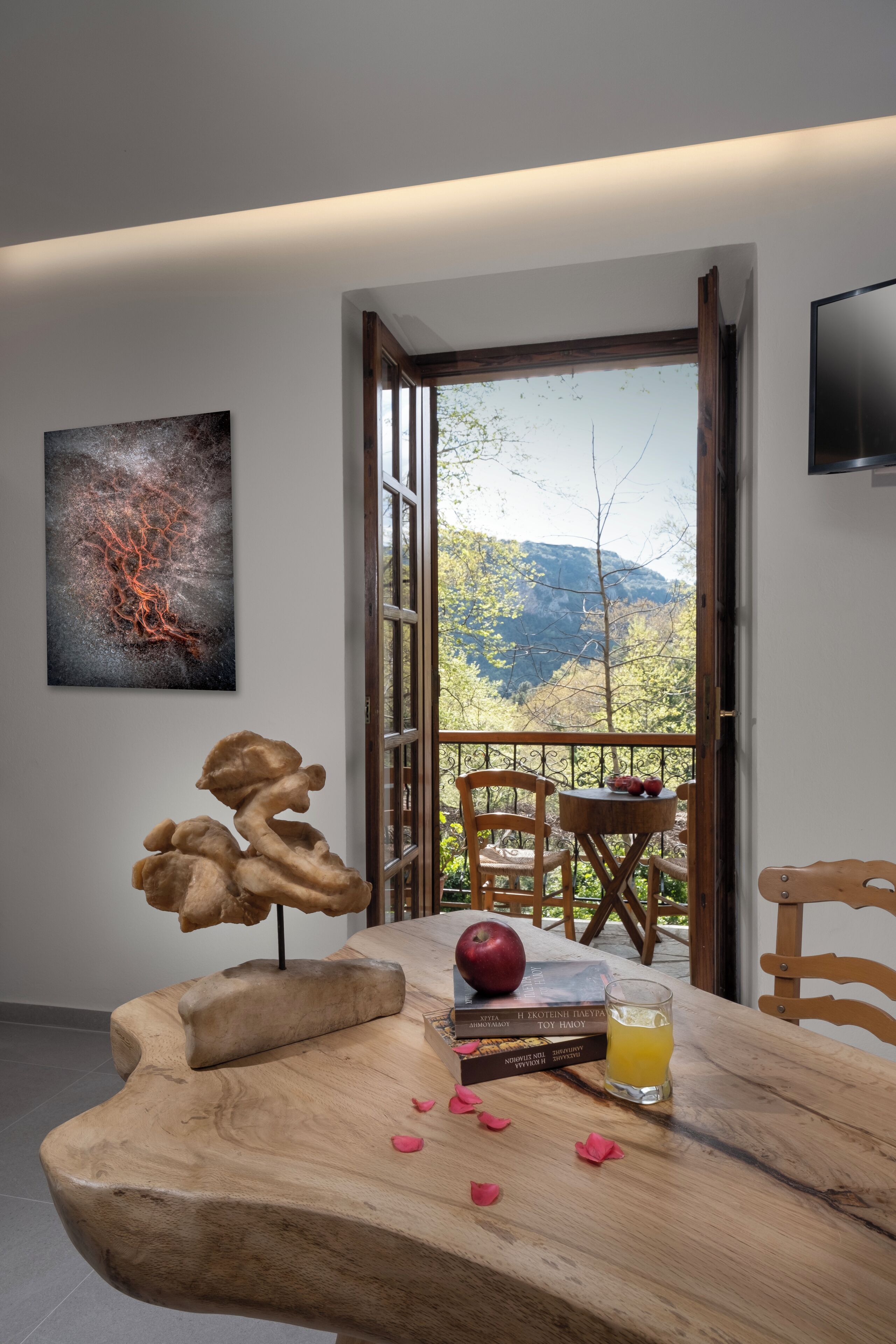 Uma mesa de madeira com um livro, uma maçã e um copo de suco. Há um quadro na parede, e uma TV fixada na parede.