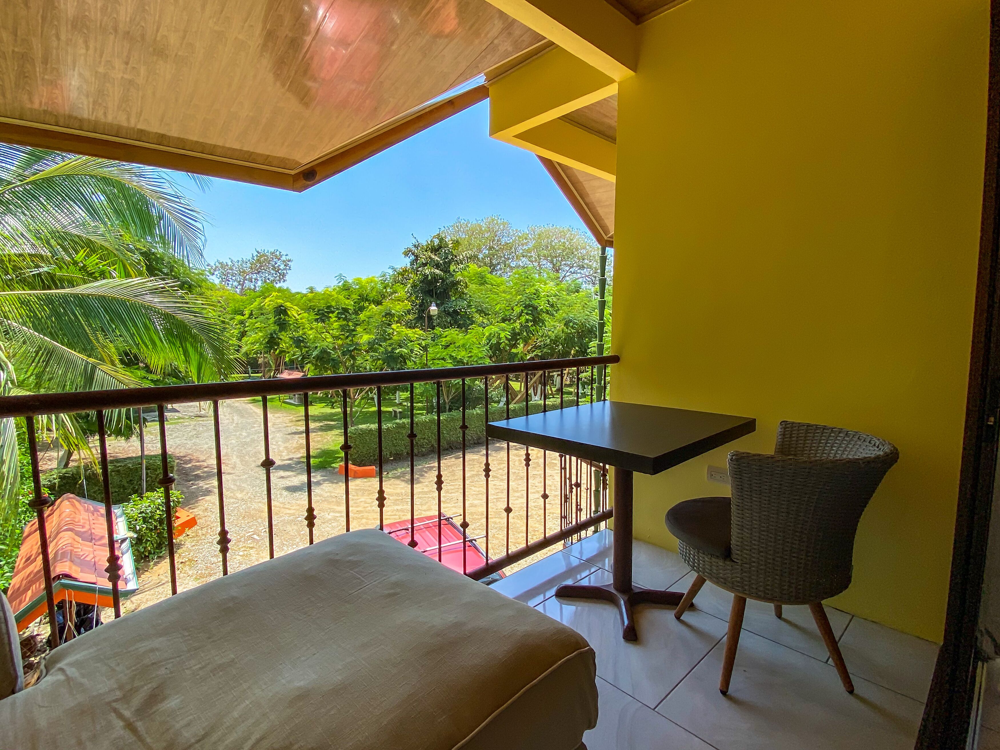 A balcony with a table and chairs, overlooking a garden with palm trees and a clear sky.