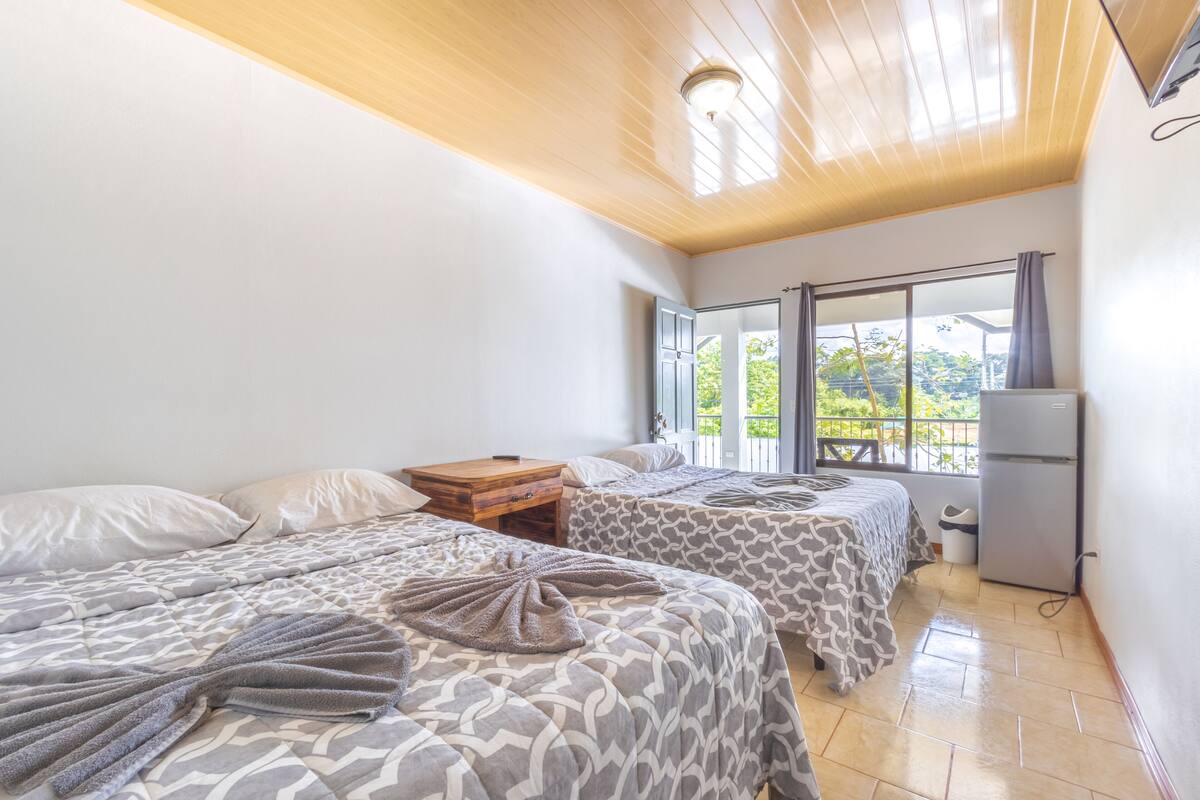 A bedroom with two beds, a wooden nightstand, a small table, a refrigerator, and a window with a view of greenery.