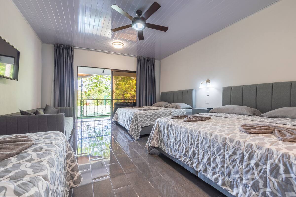 A hotel room with two beds, a ceiling fan, a large window with curtains, and a view of greenery outside.