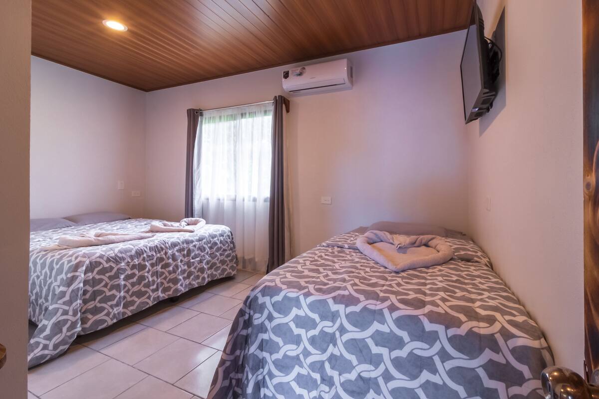 A room with two beds, each with patterned bedding, a ceiling fan, an air conditioning unit, and a television mounted on the wall.