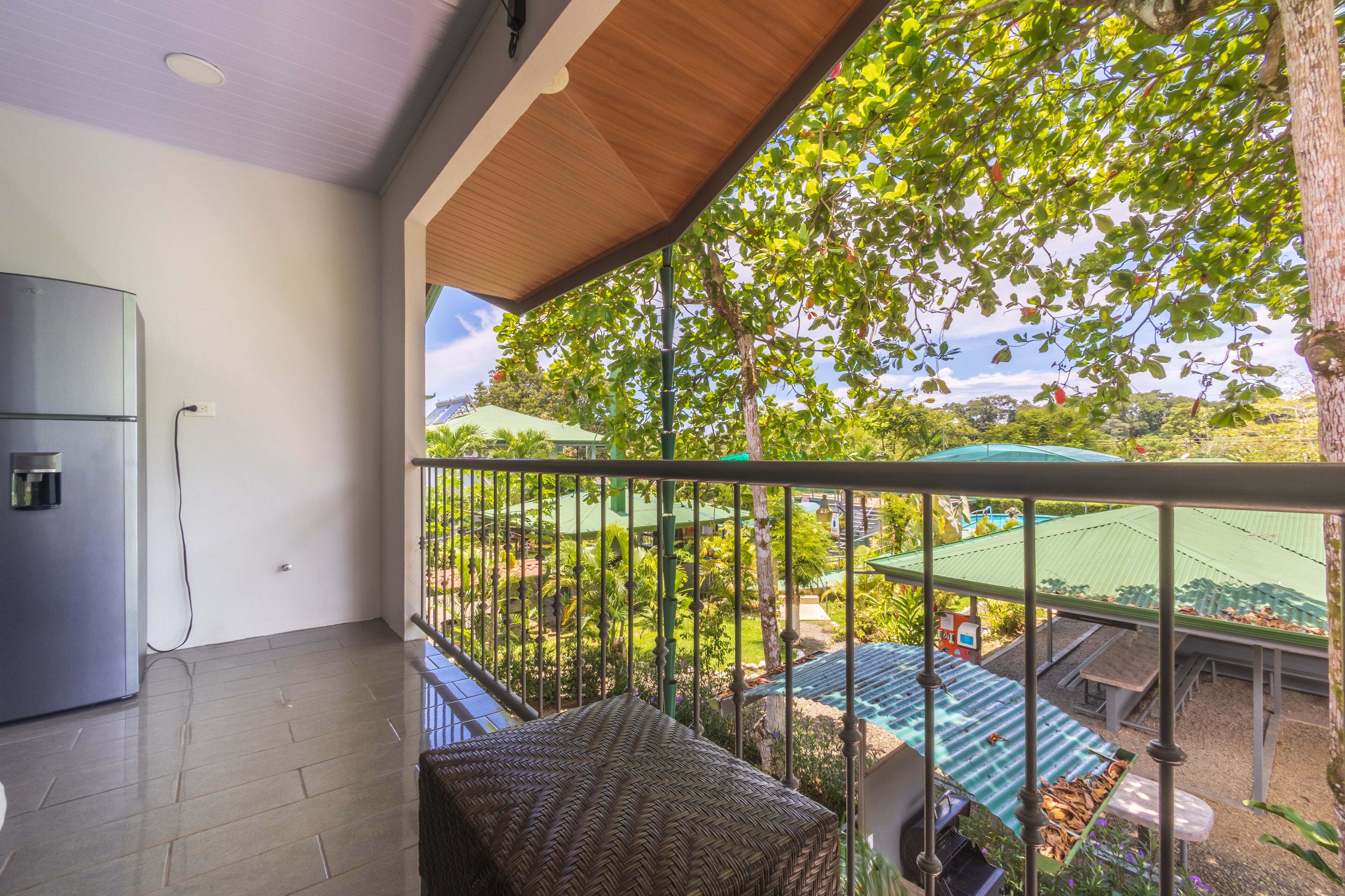 Un balcón con vista a una piscina y follaje, un refrigerador y una banca de mimbre.