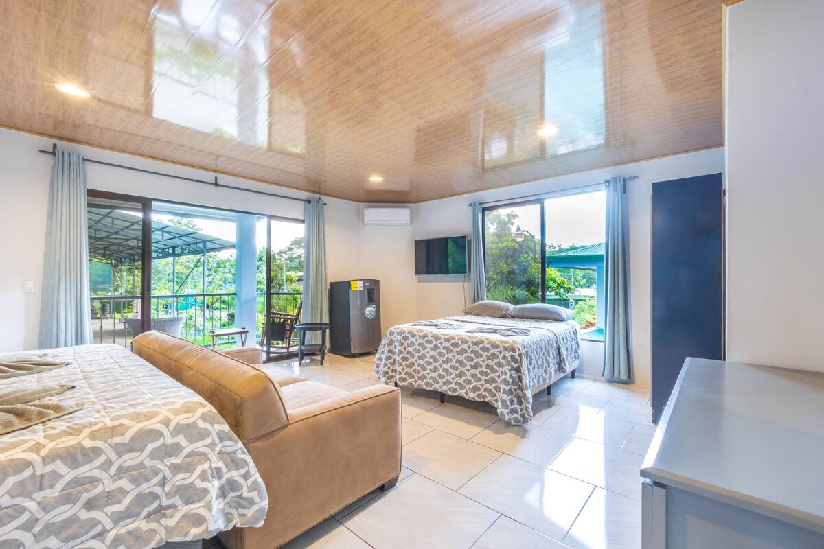A modern hotel room with two beds, a sofa, a desk, and a refrigerator. The room has large windows and a view of greenery outside.