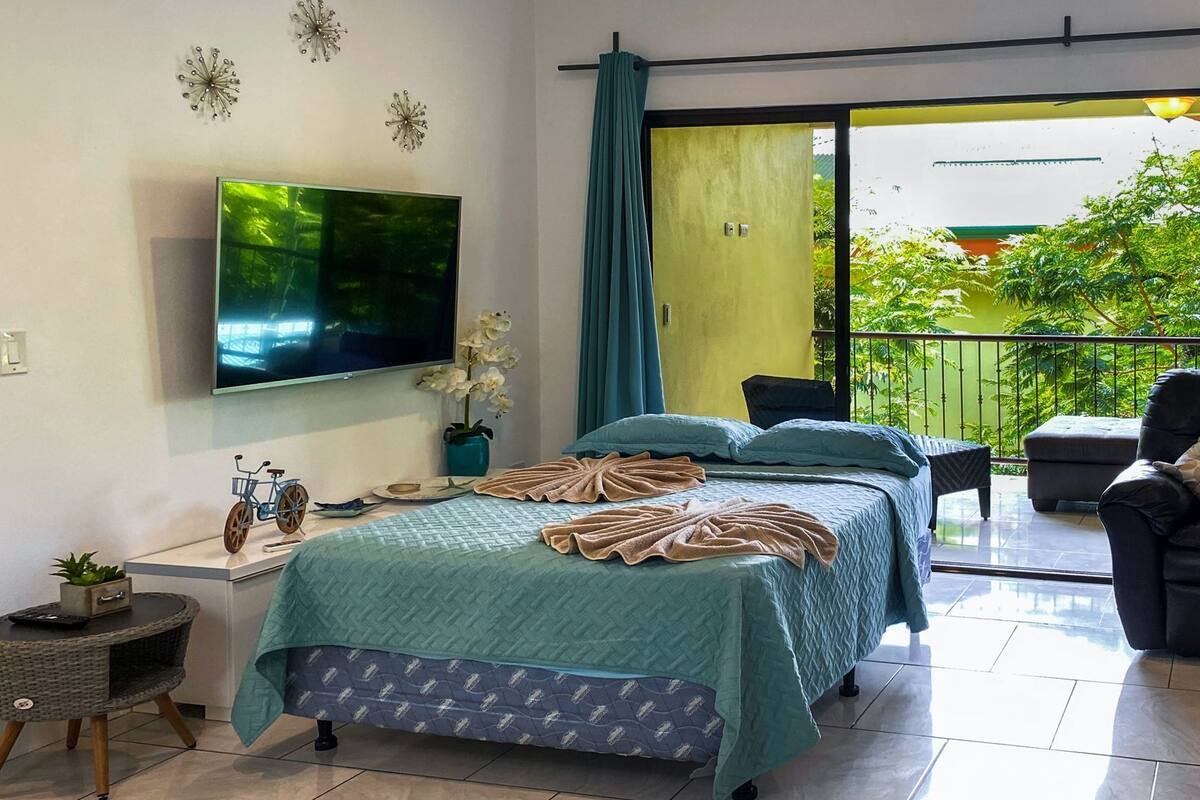 A hotel room with a bed, a TV, a small table with a plant, and a balcony with a view of greenery.