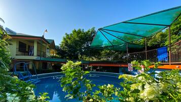 A swimming pool with a blue water surface, surrounded by lush greenery and a covered area with a green canopy.