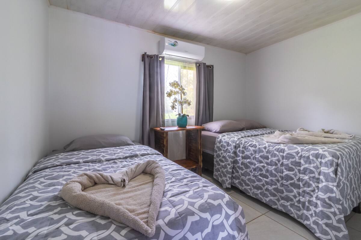 A room with two beds, a small table with a vase, and an air conditioning unit.