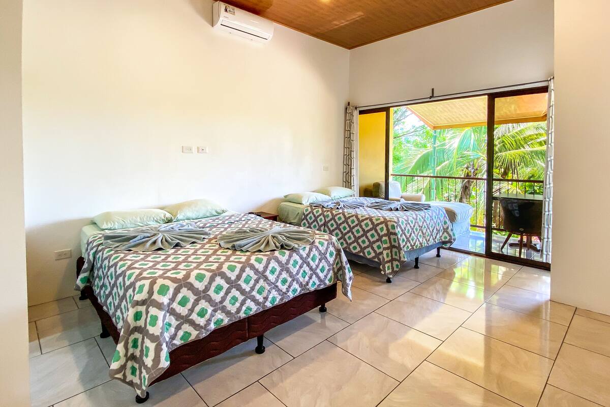 A hotel room with two beds, a large window with a view of palm trees, and a balcony with a chair.