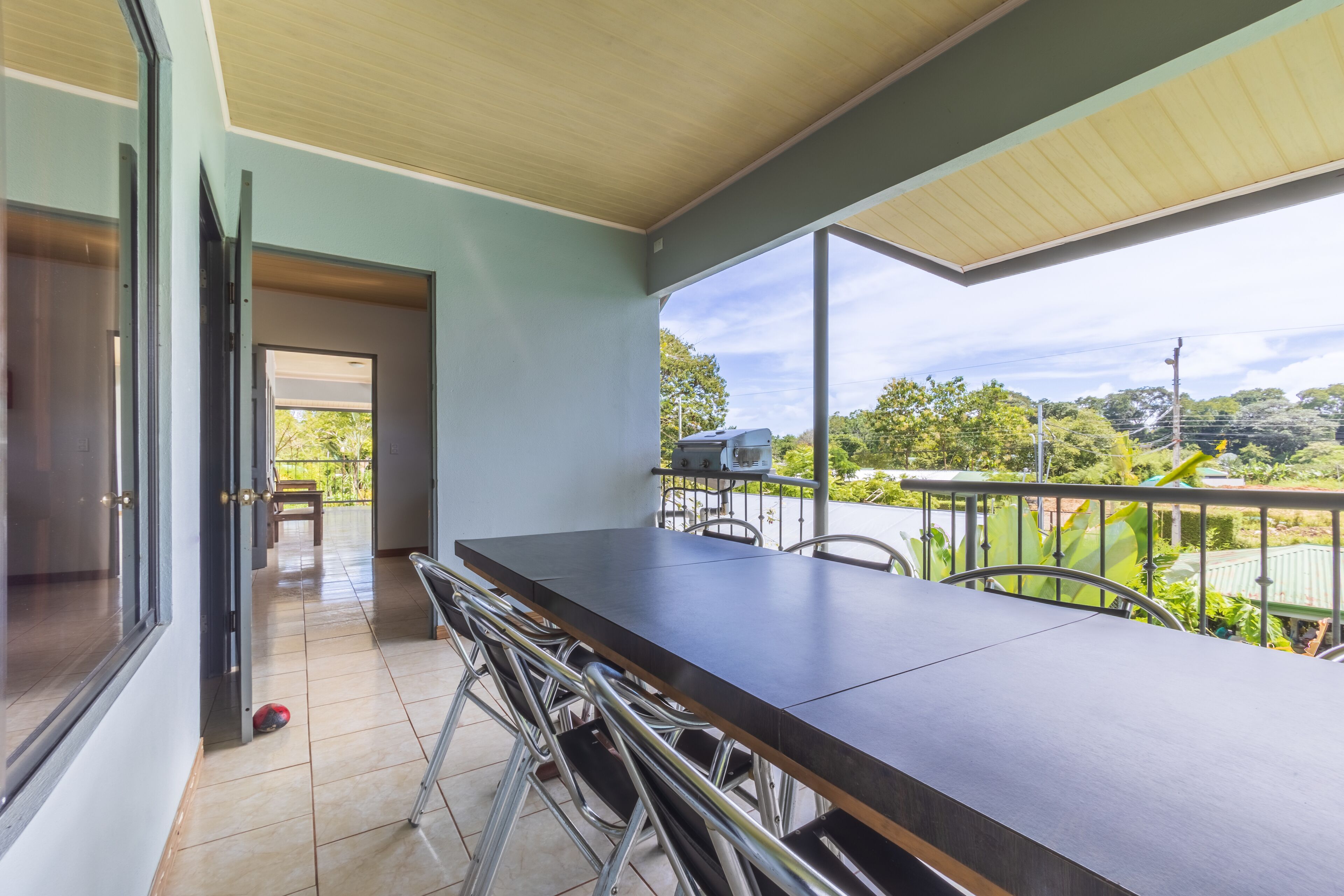 A spacious balcony with a dining table and chairs, a grill, and a view of greenery and trees.