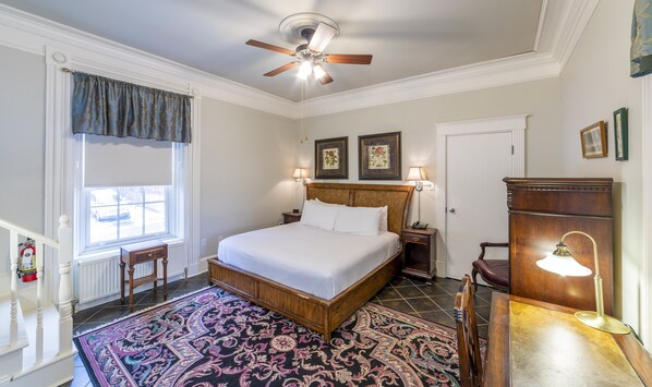 A bedroom with a large bed, a ceiling fan, a nightstand, a desk, a chair, a window with curtains, and two framed pictures on the wall.