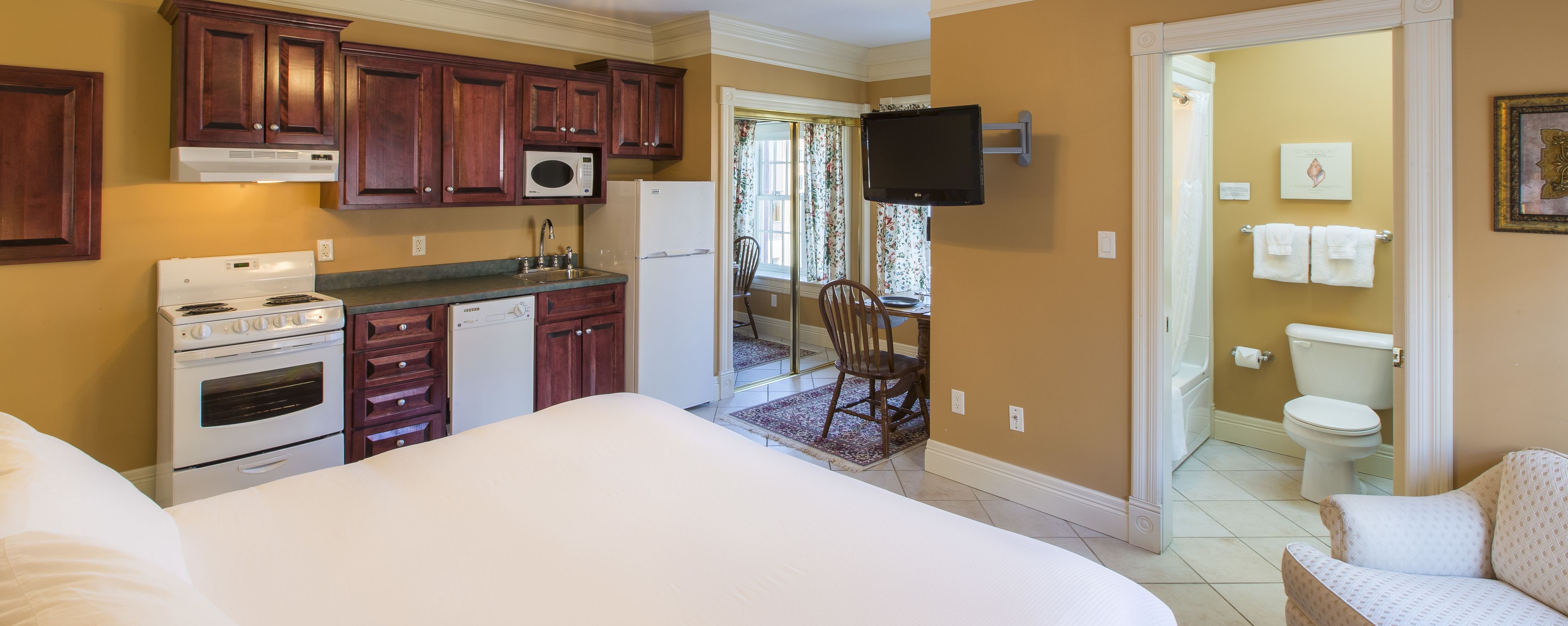 A compact hotel room with a kitchenette, a bed, a bathroom, and a TV mounted on the wall.