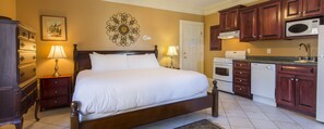 A bedroom with a large bed, wooden nightstands, a wall clock, and a kitchen area with wooden cabinets and appliances.