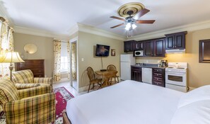 A hotel room with a bed, a dining table with chairs, a kitchen area with a microwave, stove, and refrigerator, and a ceiling fan.