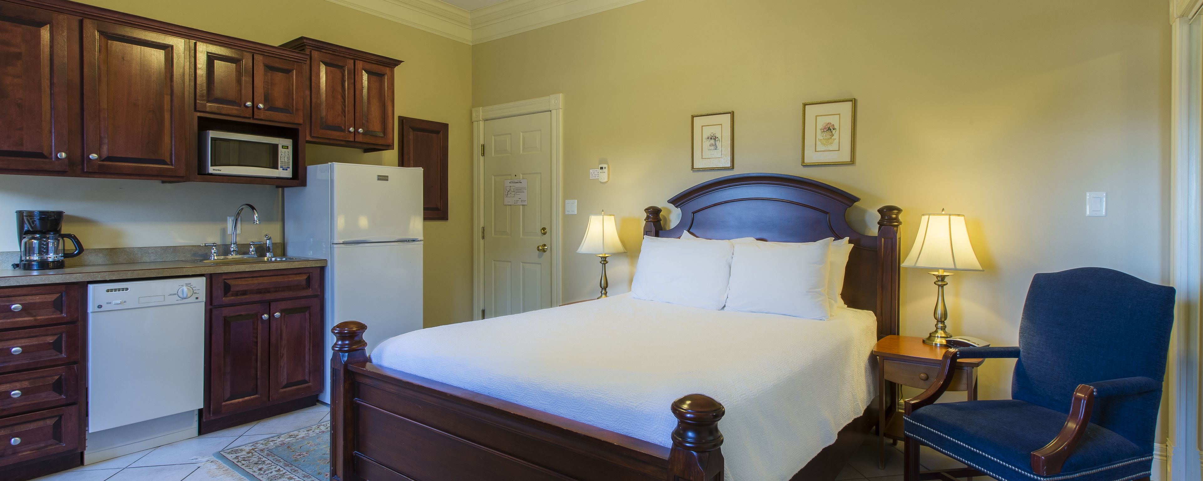 Una habitación de hotel con una cama, lámparas de noche, una silla, un escritorio, un microondas, un refrigerador y armarios de madera.