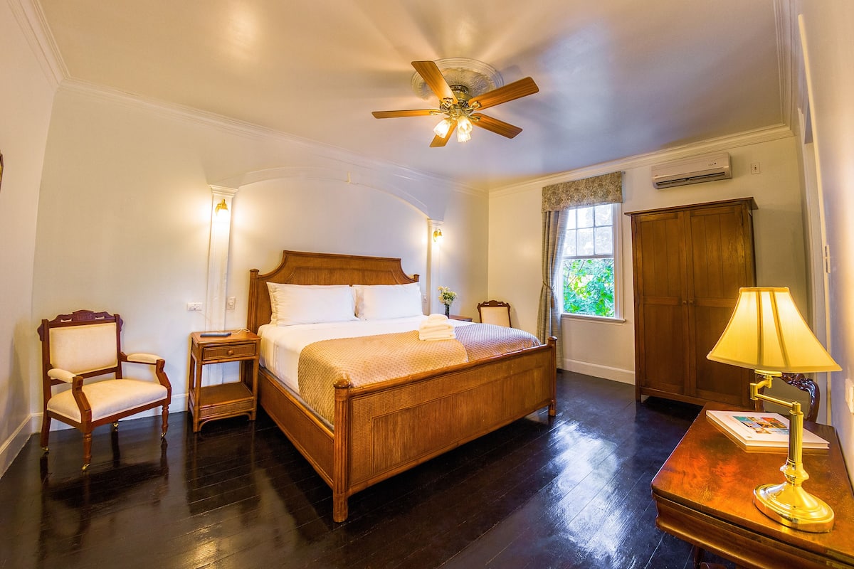 A spacious bedroom with a large wooden bed, a ceiling fan, a nightstand, a chair, a desk with a lamp, and a window with a view of greenery.