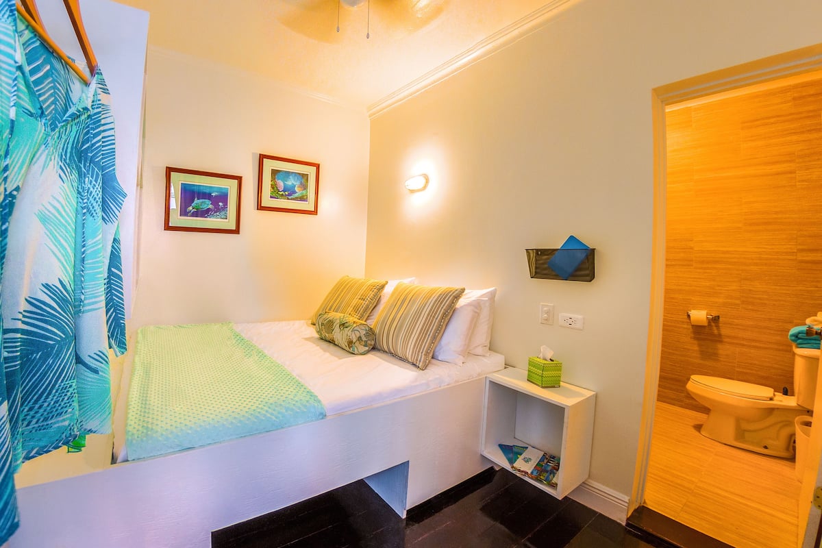 A hotel room with a bed, a bedside table with a lamp, a wall-mounted shelf with a tissue box, and a bathroom with a toilet and sink.