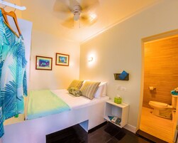 Habitación de hotel con cama, mesita de noche con lámpara, estante en la pared con caja de pañuelos y baño con inodoro y lavamanos.