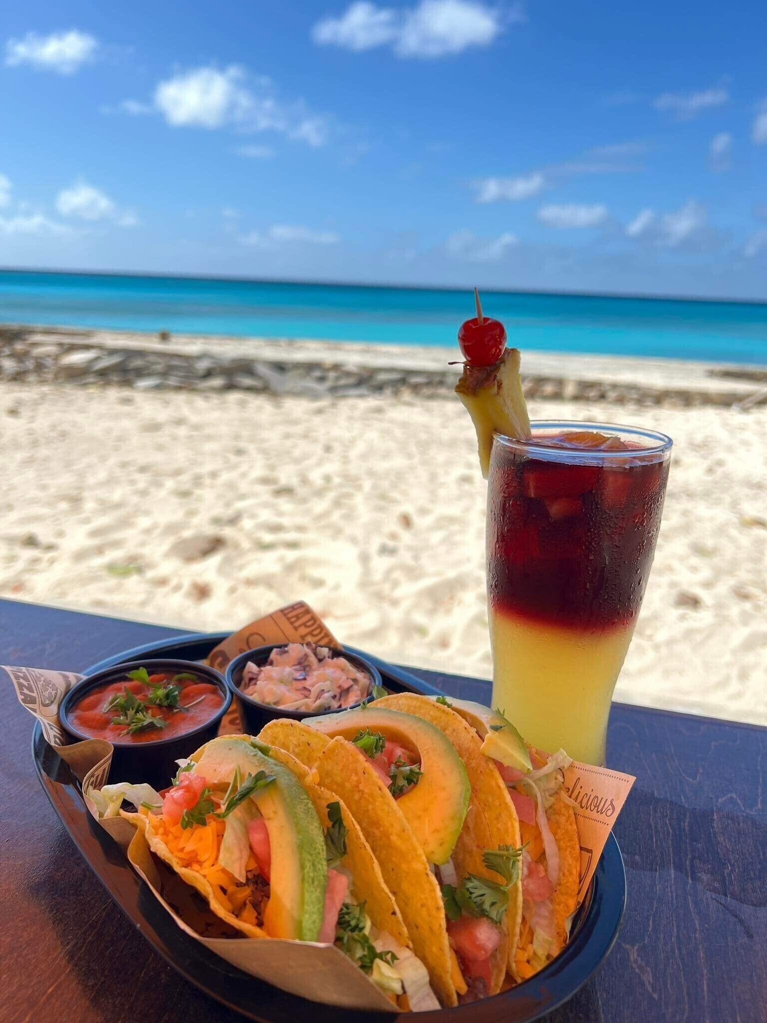 A table with tacos, a drink, and small bowls of salsa and guacamole on a beach with a clear blue sky.
