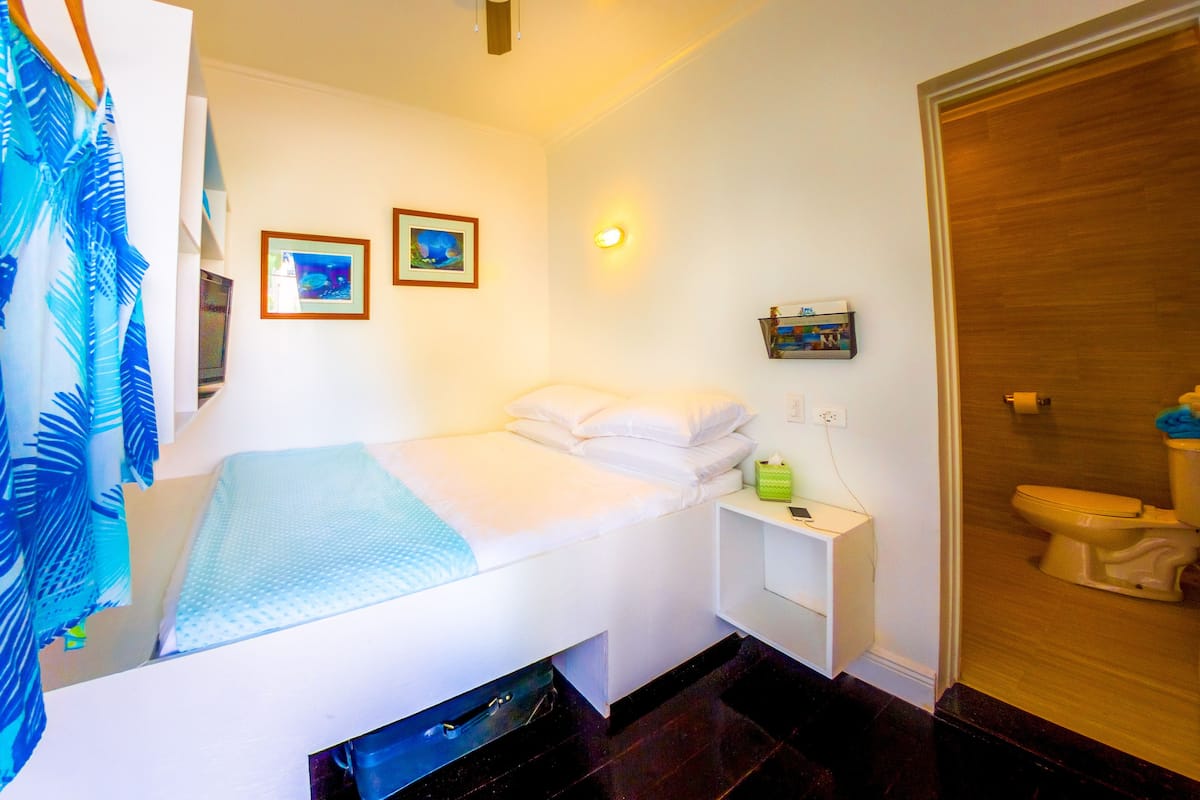 A small, single-bed room with a ceiling fan, a bedside table with a lamp, and a toilet.
