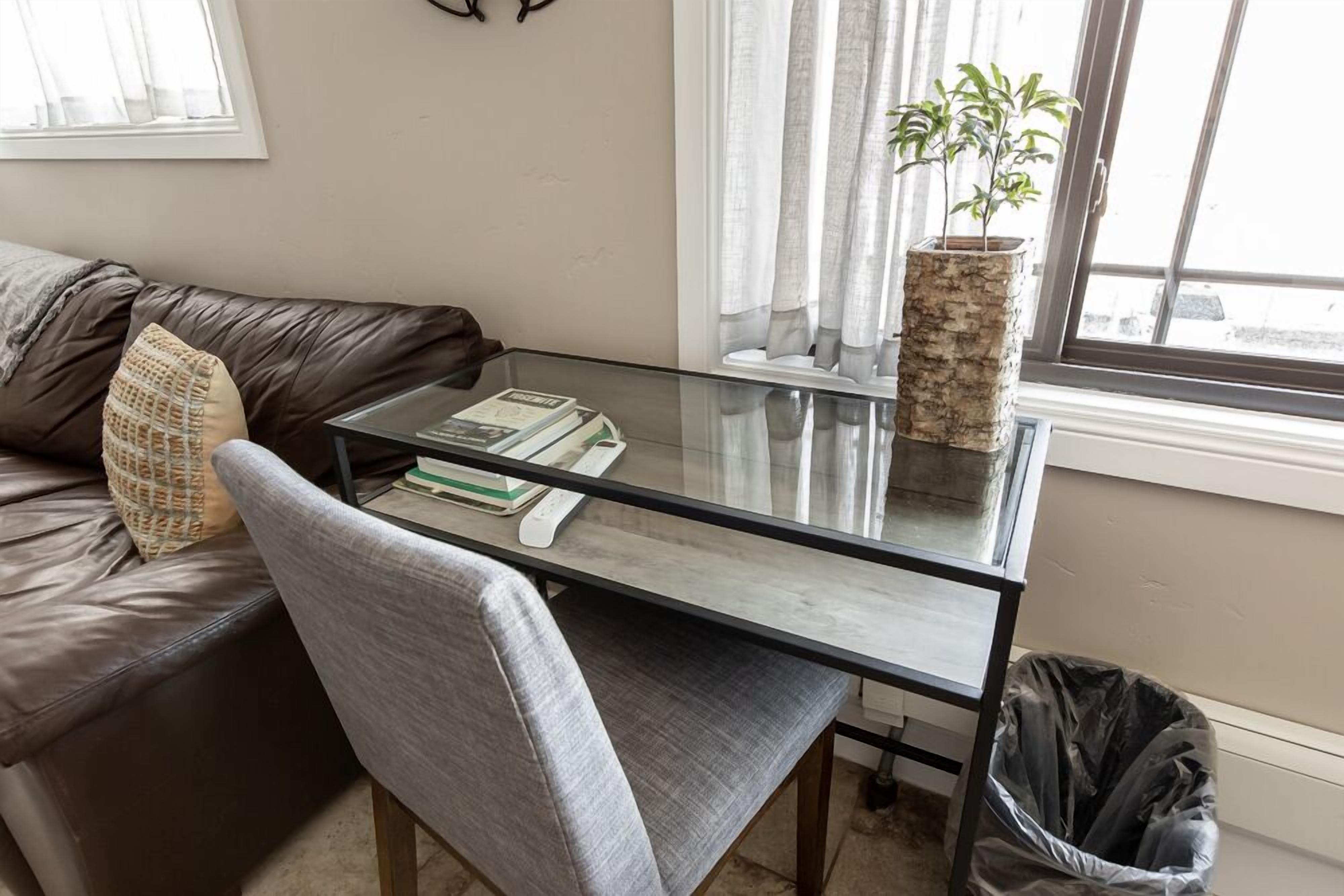 Un salón con una mesa de centro de vidrio, un sillón gris, un sofá de cuero marrón, una planta en maceta y una ventana con cortinas.