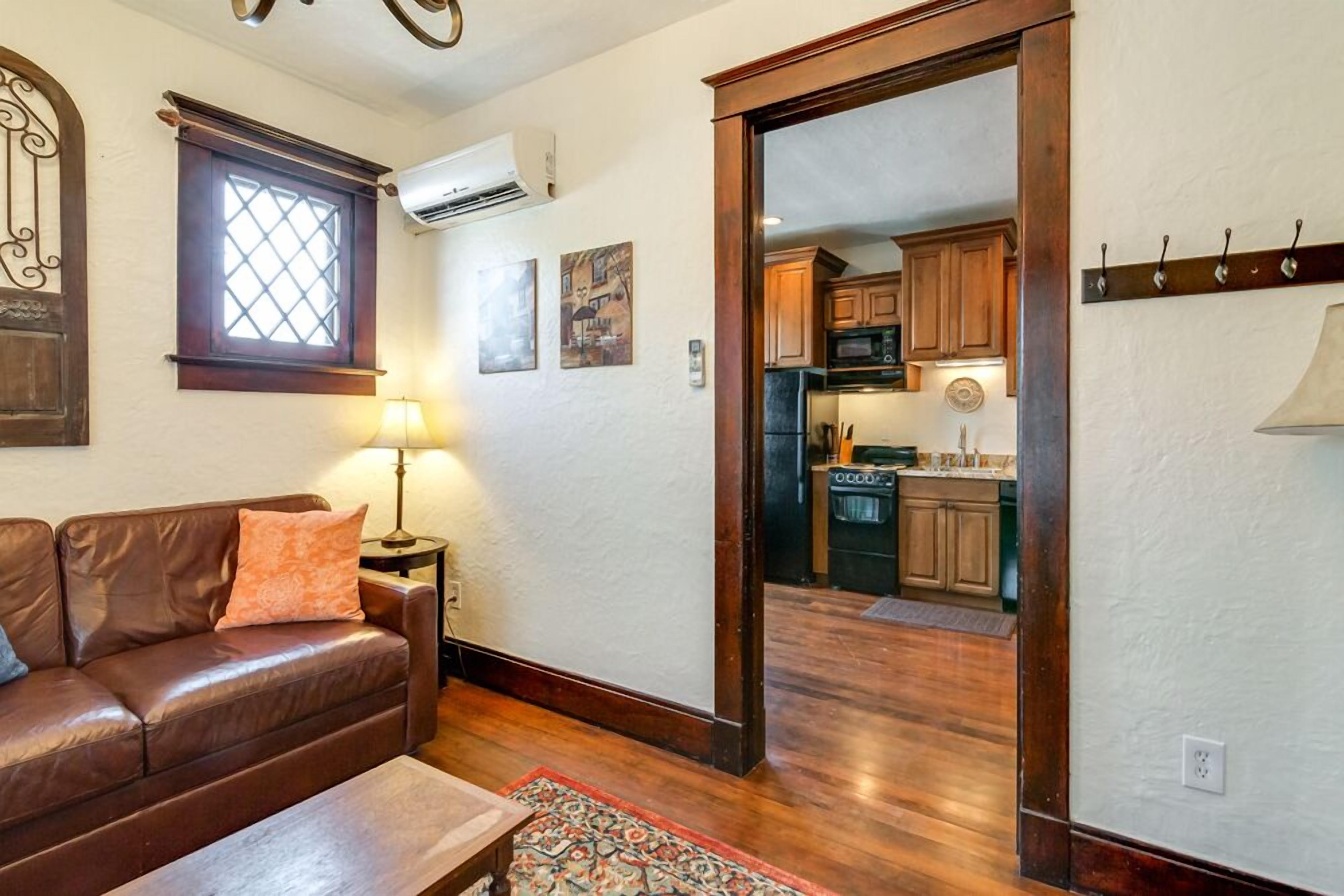 A living room with a brown leather sofa, a patterned rug, and a wooden coffee table. There is a window with a decorative frame and a wall-mounted lamp. A doorway leads to a kitchen with wooden cabinets and a microwave.