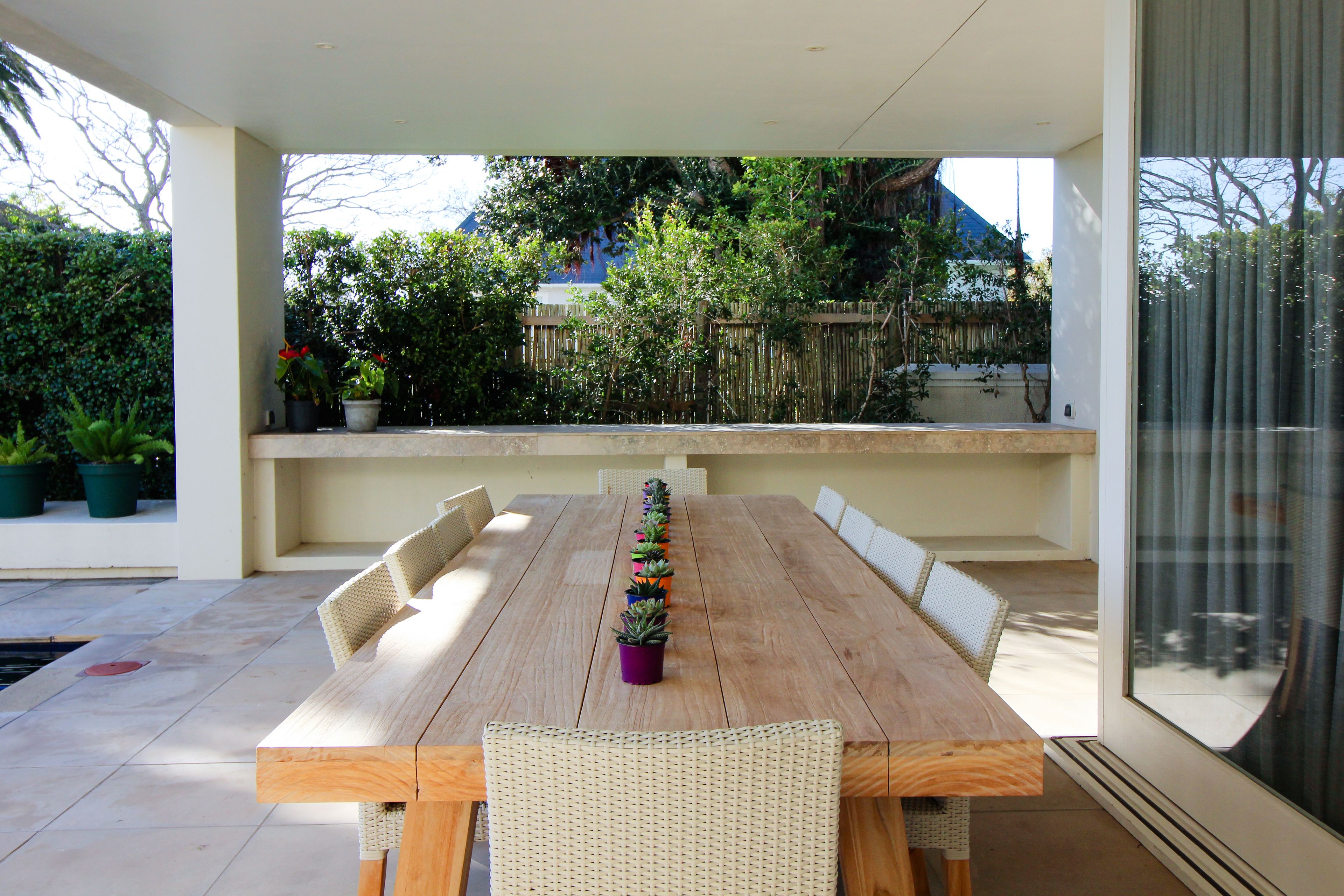 An outdoor dining area with a long wooden table and wicker chairs, surrounded by potted plants and a view of a building in the background.