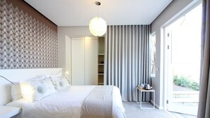 Quarto moderno com uma cama grande, mesa de cabeceira com chaleira, guarda-roupa e vista para o exterior através de uma porta deslizante.