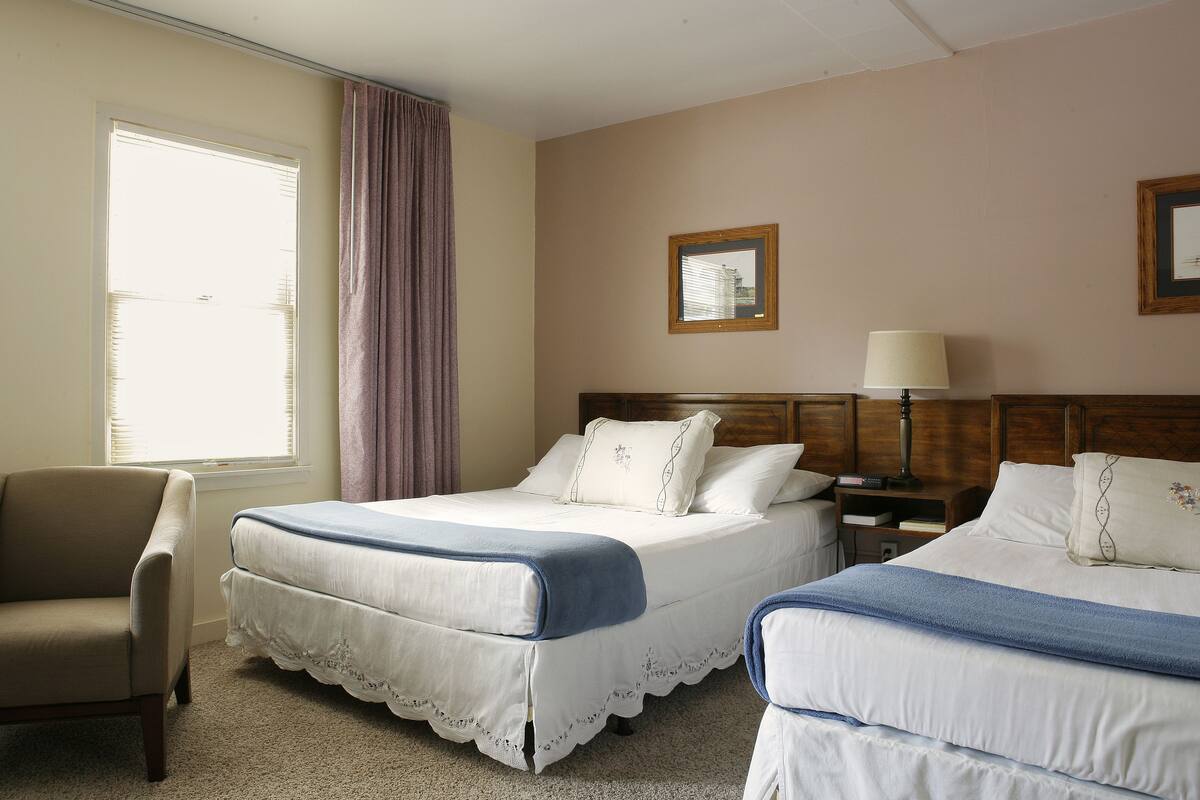 A hotel room with two beds, a chair, a nightstand with a lamp, and a window with curtains.