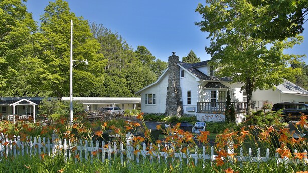 Une maison avec une cheminée en pierre, entourée d’une clôture en bois peinte en blanc et agrémentée de fleurs orange.