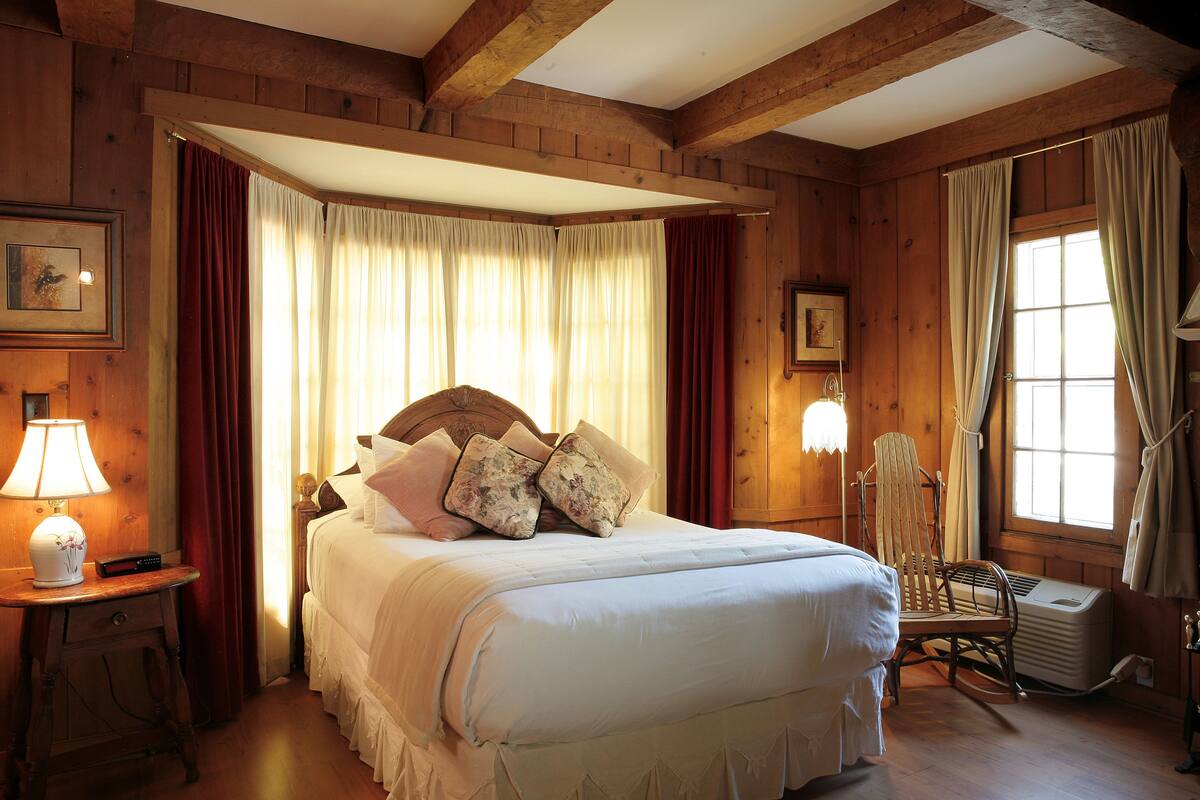A wooden room with a bed, a chair, a lamp, and a window with curtains.