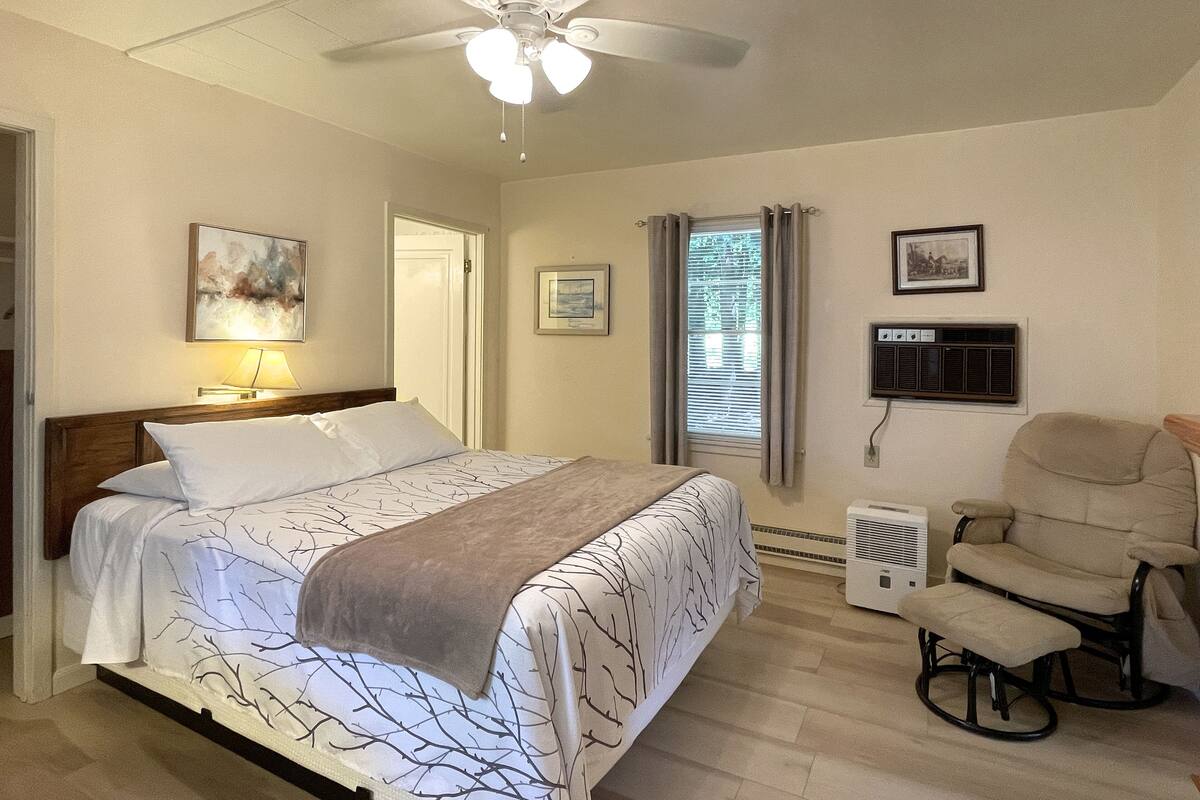 A bedroom with a bed, a chair, a ceiling fan, a window with curtains, and wall-mounted air conditioning unit.