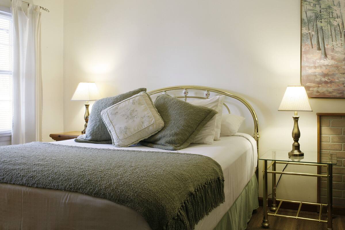 A neatly made bed with a green blanket, two bedside lamps, and a painting on the wall.