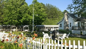 Une maison blanche avec une cheminée en pierre, entourée d’une clôture en bois peinte en blanc, et un kiosque dans la cour.
