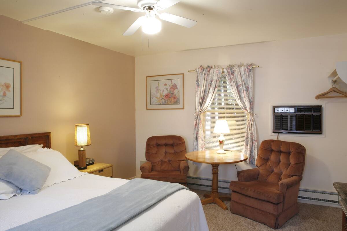 A hotel room with a bed, two armchairs, a small table, a ceiling fan, a window with curtains, and a wall-mounted air conditioning unit.