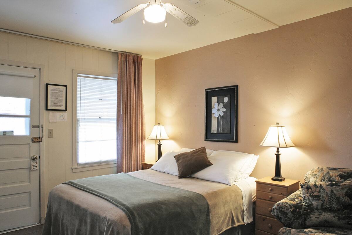 A hotel room with a bed, two bedside lamps, a ceiling fan, a framed picture on the wall, a window with curtains, and a chair.